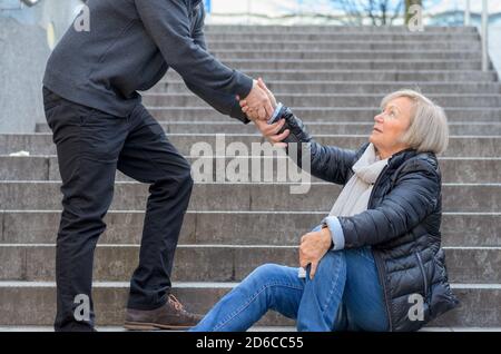 Mann hilft senior Frau aufzustehen, während sie auf Treppen im Freien in der Stadt sitzt, halten Sie ihr Knie Stockfoto