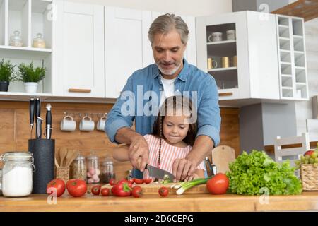 Nettes kleines Mädchen und ihr hübscher Vater schneiden Gemüse und lächeln beim Kochen in der Küche zu Hause. Stockfoto