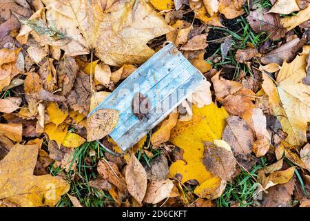 Schmutzige gebrauchte Sanitär-Gesichtsmaske in nassen abgefallenen Blättern auf Rasen im Stadtpark im Herbst Stockfoto