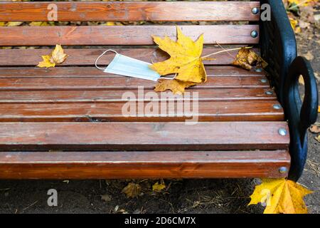 Fallengelassene medizinische Gesicht fask und gelbe Ahornblätter auf Holz Bank im Stadtpark am Herbsttag Stockfoto