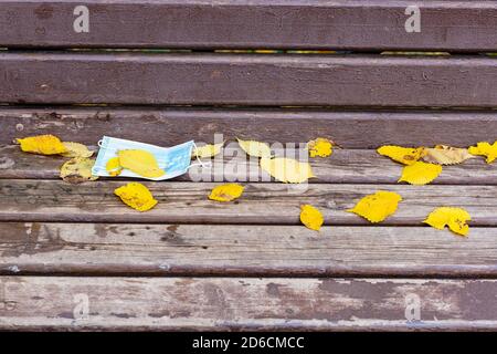 Fallengelassene Sanitär-Gesichtsmaske mit gelb gestürzten Blättern bedeckt auf Schäbige Holzbank im Stadtpark am Herbsttag Stockfoto