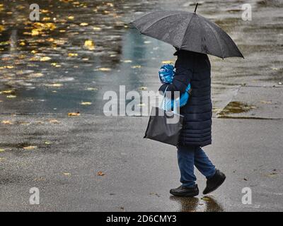 Person mit Sonnenschirm geht durch den Regen. Person mit Sonnenschirm geht über eine nasse Straße. Person mit Regenschirm im Regen. Der Herbst hinterlässt auf dem We Stockfoto