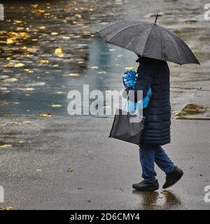 Person mit Sonnenschirm geht durch den Regen. Person mit Sonnenschirm geht über eine nasse Straße. Person mit Regenschirm im Regen. Der Herbst hinterlässt auf dem We Stockfoto