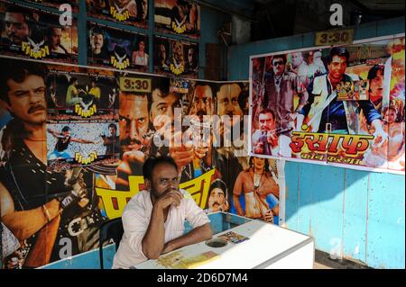 13.12.2011, Mumbai, Maharashtra, Indien - EIN Mann sitzt an der Kasse in einem kleinen Kino mit bunten Filmplakaten im Dharavi-Slum und isst ein Eis Stockfoto