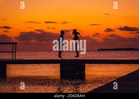 Silhouetten eines Jungen und eines Mädchens, die im Abenduntergang in den Florida Keys auf einem Dock springen. Stockfoto