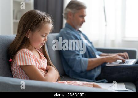 Keine Zeit Für Kind. Grauhaarige Papa beschäftigt mit Laptop, online zu Hause arbeiten, traurig gelangweilt beleidigt Tochter sitzt in der Nähe Stockfoto