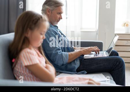 Keine Zeit Für Kind. Grauhaarige Papa beschäftigt mit Laptop, online zu Hause arbeiten, traurig gelangweilt beleidigt Tochter sitzt in der Nähe Stockfoto