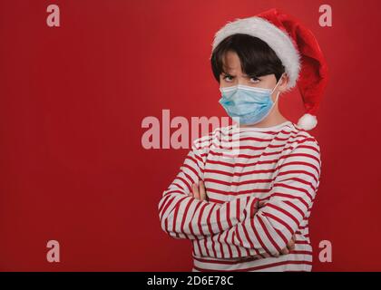 Wütend Kind mit medizinischen Maske trägt Weihnachtsmann Hut über Roter Hintergrund Stockfoto