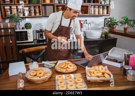 Ein professioneller männlicher Koch mit einem Handy bereitet ein Donut Auftrag Halten Sie ein digitales Tablet in der Küche Stockfoto