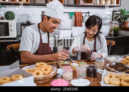 Ein Ehepaar, das in Schürzen sitzt, während er Donuts macht Und Donuts in der Küche mit Schokolade dekorieren Stockfoto