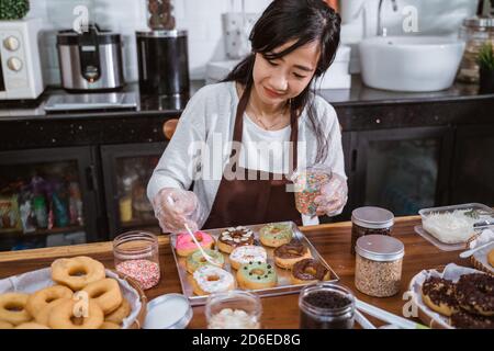 Die Köchin trägt eine Schürze, während sie Donuts mit streut Schokolade und schmückt Donuts in der Küche Stockfoto