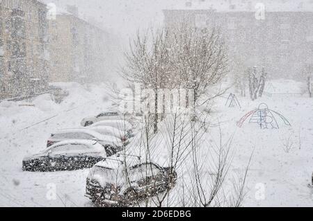 Starker Schneesturm in der Stadt. Autos mit Schnee bedeckt auf Parkplatz in Wohngebiet während Dezember Schneesturm. Winterliche Stadtlandschaft bei Schneefall. Winterwein Stockfoto