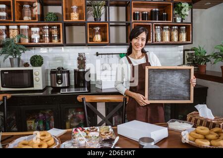 Eine attraktive Köchin trägt eine Schürze mit einem schwarzen Tafel, während man im Küchenraum steht Stockfoto