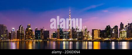 Atemberaubender Blick auf die beleuchtete Skyline von Dubai in der Abenddämmerung mit modernen Wolkenkratzern, die sich auf dem im Vordergrund fließenden Wasserkanal spiegeln. Stockfoto