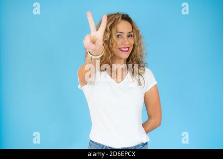 Blonde Frau mit lockigem Haar trägt Casual Shirt lächeln mit glücklichem Gesicht zwinkert an der Kamera tun Siegeszeichen. Nummer zwei. Stockfoto