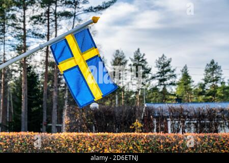 Nahaufnahme der schwedischen Flagge mit Fahnenmast, der bei ruhigem Wetter langsam winkt. Verschwommen schön gefärbt gut getrimmt Herbst Hecken, Kiefernwald und Stockfoto