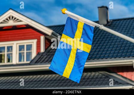 Nahaufnahme der schwedischen Flagge, verschwommenes schwarzes Dach und Fenster oben auf dem schwedischen roten Holzhaus im Hintergrund. Blauer Himmel an sonnigen Tagen Stockfoto