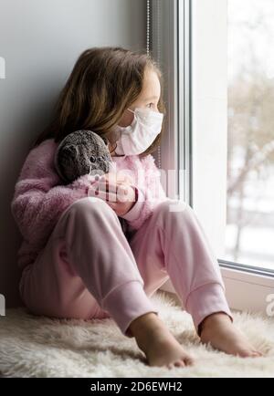 COVID-19 Coronavirus-Konzept, trauriges Kind in medizinischer Maske schaut aus Fenster im Haus. Nachdenkliches Kind sitzt im Winter zu Hause auf der Fensterbank. Kleines Mädchen mit Stockfoto