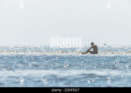 Mann in Badehose sitzt im Wasser im Wattenmeer und beobachtet die Wellen und die Flut. Stockfoto