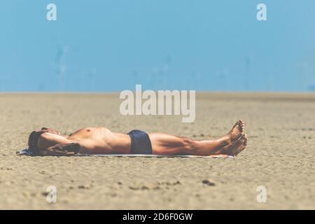 Mann in Badehose liegt im Sand und Sonnenbaden, St. Peter Ording, Strandurlaub, Windturbinen im Hintergrund Stockfoto