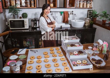 Selbstbewusster Koch asiatische Frau, die in einer modernen Küche lächelt Mit gekreuzten Händen vor Donuts auf dem Tisch In der Küche Stockfoto