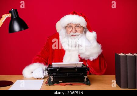 Foto von santa claus grauen Bart sitzen Schreibtisch Faust Wange Vintage Schreibmaschine Lampe Papier Buch tragen x-Mas Kostüm Mantel Mütze Gläser isoliert rote Farbe Stockfoto