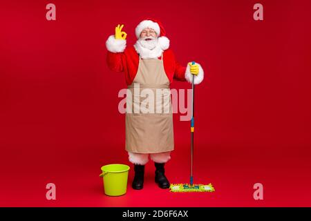 In voller Länge Foto von santa claus grauen Bart halten Mopp Zeige Okey Wear x-Mas Kostüm Schürze Mantel Kopfbedeckung Brille Latex Handschuhe Stiefel Brille isoliert rot Stockfoto