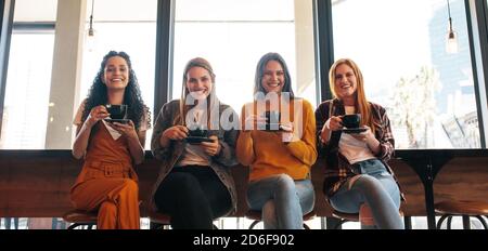 Porträt einer glücklichen jungen Freundinnen, die in einem Café zusammen sitzen und Kaffeetassen halten und die Kamera betrachten. Eine Gruppe von Frauen trifft sich in einem Café Stockfoto