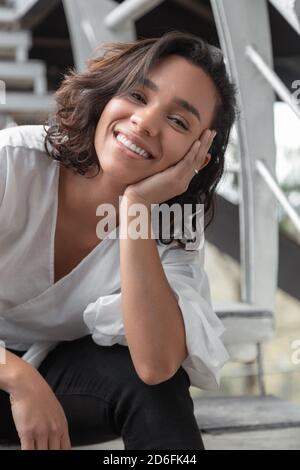 Fröhliche junge lateinische Brünette Frau mit Locken und kurzen Haaren  sitzt auf der Treppe, lächelnd, trägt weiße Bluse und schwarze Hose, Foto  tagsüber Stockfotografie - Alamy