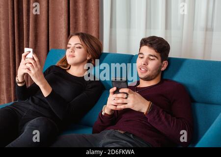 Fröhliches Paar, das Nachrichten auf Handys schreibt, sitzt auf einer blauen Couch zu Hause. Konzept für Social Media-Chats. Stockfoto