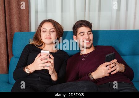 Fröhliches Paar, das Nachrichten auf Handys schreibt, sitzt auf einer blauen Couch zu Hause. Konzept für Social Media-Chats. Stockfoto