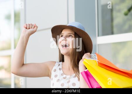 Aufgeregt junge Dame mit Bündel von Papiertüten gestikulieren mit Hand und Blick nach oben, während Sie auf der Stadtstraße stehen Sonniger Tag Stockfoto