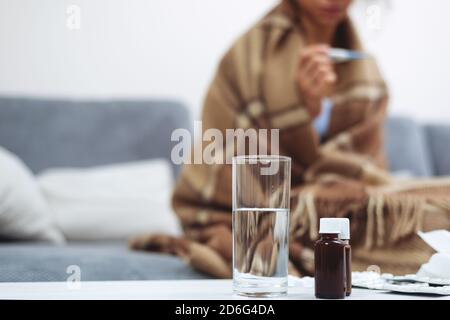 Stillleben von Medikamenten auf dem Nachttisch. Im Hintergrund die junge Frau mit einem Thermometer in den Händen. Behandlung zu Hause. Hohe Körpertemperatur Stockfoto