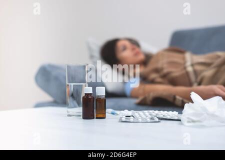 Stillleben von Medikamenten auf dem Nachttisch. Im Hintergrund liegt die kranke junge Frau auf einem Bett. Behandlung zu Hause. Hohe Körpertemperatur. Pandemi Stockfoto