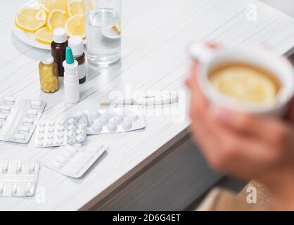 Stillleben von Medikamenten für die Behandlung der Virusinfektion zu Hause. Hände mit einer Tasse heißen Tee mit Zitrone sind sichtbar. Pandemie, Selbstisolierung. Stockfoto