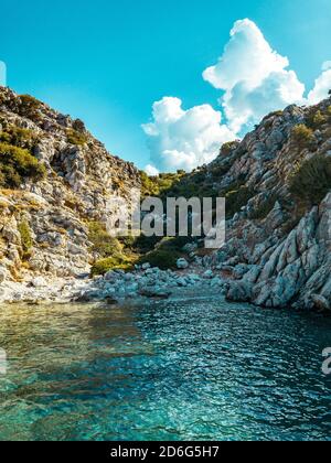 Die Sonne scheint über einer felsigen Klippe und wirft seine Schatten auf eine natürliche Meeresbucht. Kristallklares türkisfarbenes Wasser und blauer Himmel mit weißen Wolken. Stockfoto