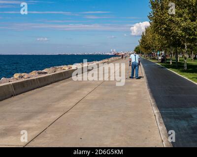 Istanbul / Türkei - 10.1.2020; ein älterer Mann läuft an einem sonnigen Tag mit Krücken auf einem von Bäumen umgebenen Parkpflaster am Meer. Stockfoto