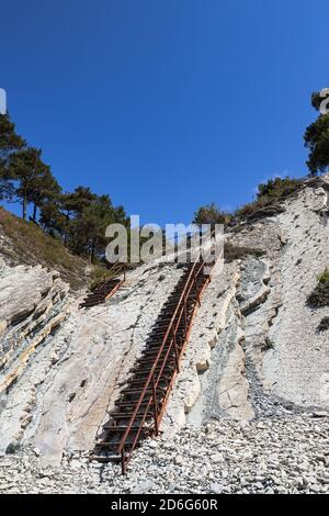 Eine alte Metalltreppe und massive weiße Felsen an einem wilden Strand. Der Weg zum Campingplatz führt durch den Wald. Sommerlandschaft an einem klaren Tag Stockfoto