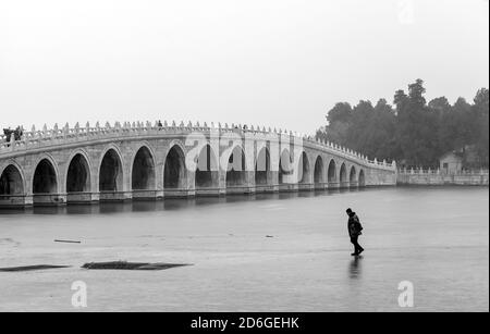 Ein Mann, der auf dem Eis in der Nähe der Brücke des 17. Arches (Shiqikong Qiao) am Kunming-See in Peking, China, läuft. Ein nebliger Wintertag Stockfoto