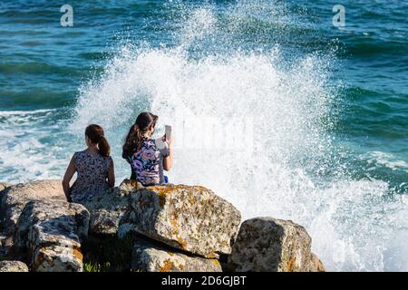 PORTONOVO, SPANIEN - 21. AUGUST 2020: Eine junge Frau macht ein Foto mit ihrem Smartphone, während sie den Sonnenuntergang auf den Felsen im Rias Baixas, Galic, genießt Stockfoto