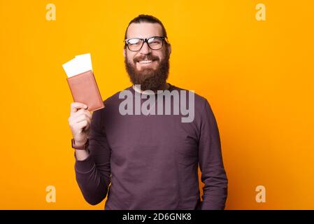 Bärtiger Mann lächelt an der Kamera und hält einen Pass mit zwei Tickets in der Nähe einer gelben Wand. Stockfoto