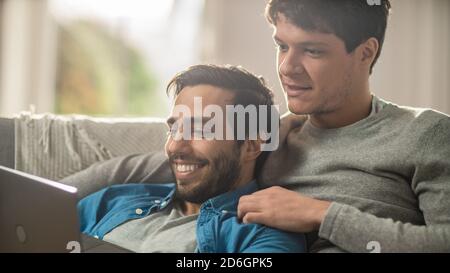 Süßes männliches Queer-Paar verbringt Zeit zu Hause. Sie liegen auf einem Sofa und benutzen den Laptop. Sie Durchsuchen Online. Die Hand des Partners ist um seinen Liebhaber herum Stockfoto