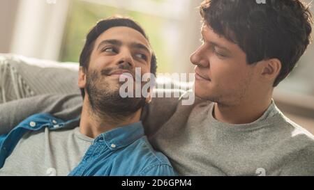 Süßes männliches Queer-Paar verbringt Zeit zu Hause. Sie liegen auf einem Sofa und schauen sich gegenseitig an. Die Hand des Partners ist um seinen Liebhaber herum. Sie sind es Stockfoto