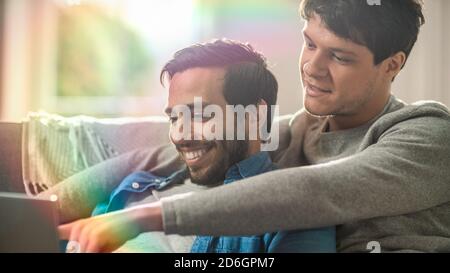 Männliches Queer-Paar verbringt Zeit zu Hause. Sie liegen auf dem Sofa und benutzen einen Laptop. Sie Durchsuchen Online. Die Hand des Partners ist um seinen Liebhaber herum und zeigt Stockfoto