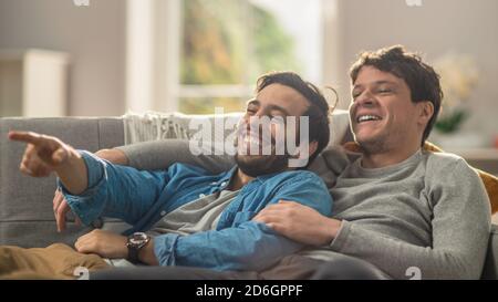 Süßes männliches Queer-Paar verbringt Zeit zu Hause. Sie liegen auf einem Sofa und lachen. Die Hand des Partners ist um seinen Liebhaber herum und zeigt auf Stockfoto