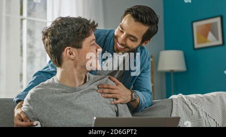 Süß Männlich Gay Paar verbringen Zeit bei zu Hause. Der junge Mann arbeitet auf einem Laptop, sein Partner kommt von hinten und umarmt ihn sanft. Sie Lachen. Das Zimmer Hat Stockfoto