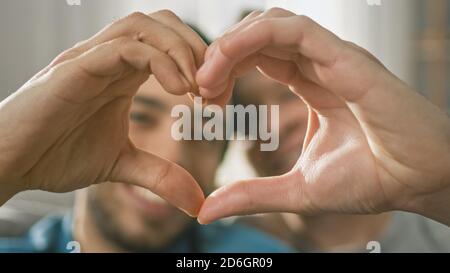 Porträt eines niedlichen männlichen Queer Paar zu Hause sitzen auf einem Sofaund machen ein Herz-Symbol mit ihren Händen. Das Zimmer Ist Modern Eingerichtet. Stockfoto