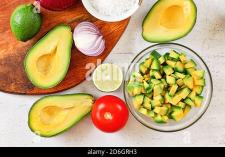 Avocado Hälften, einige in kleine Stücke geschnitten, Limetten, Tomaten und Zwiebeln - Grundzutaten Guacamole auf weißem Brett, Blick von oben Stockfoto