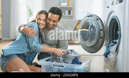Schöne junge Paar sitzen neben einer Waschmaschine zu Hause. Sie lachen und umarmen, während sie die Waschmaschine mit schmutziger Wäsche beladen. Hell und geräumig Stockfoto