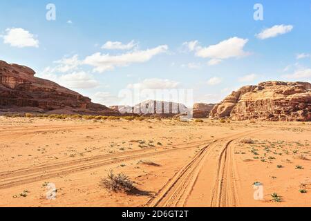 Felsige Massive auf roter Sandwüste, Fahrzeugspuren Boden, heller wolkiger Himmel im Hintergrund, typische Landschaft im Wadi Rum, Jordanien Stockfoto